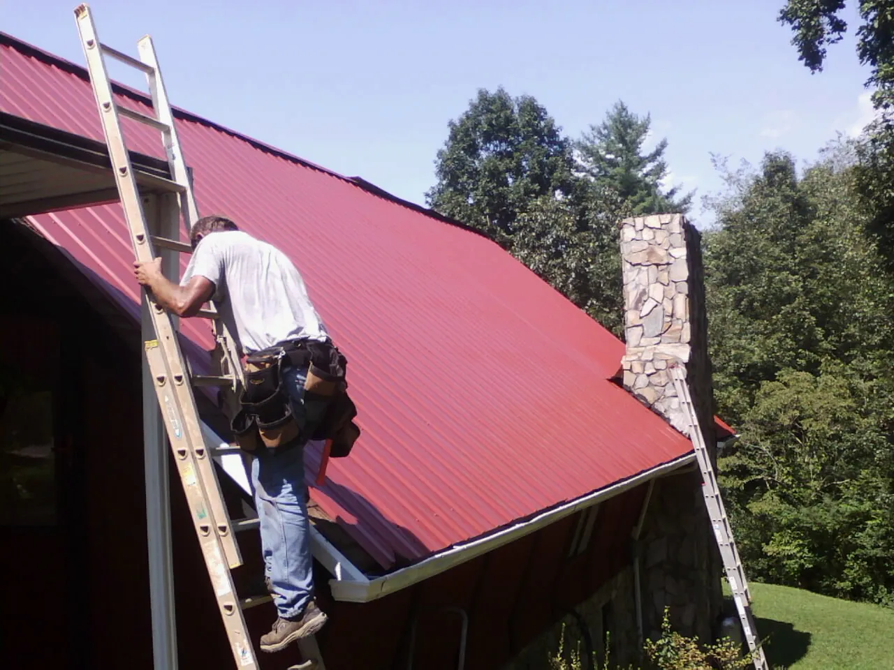 Roofer on a ladder beside a red metal roof and stone chimney — Soffit & Fascia Repair by Roofing Brothers in Cambridge Springs